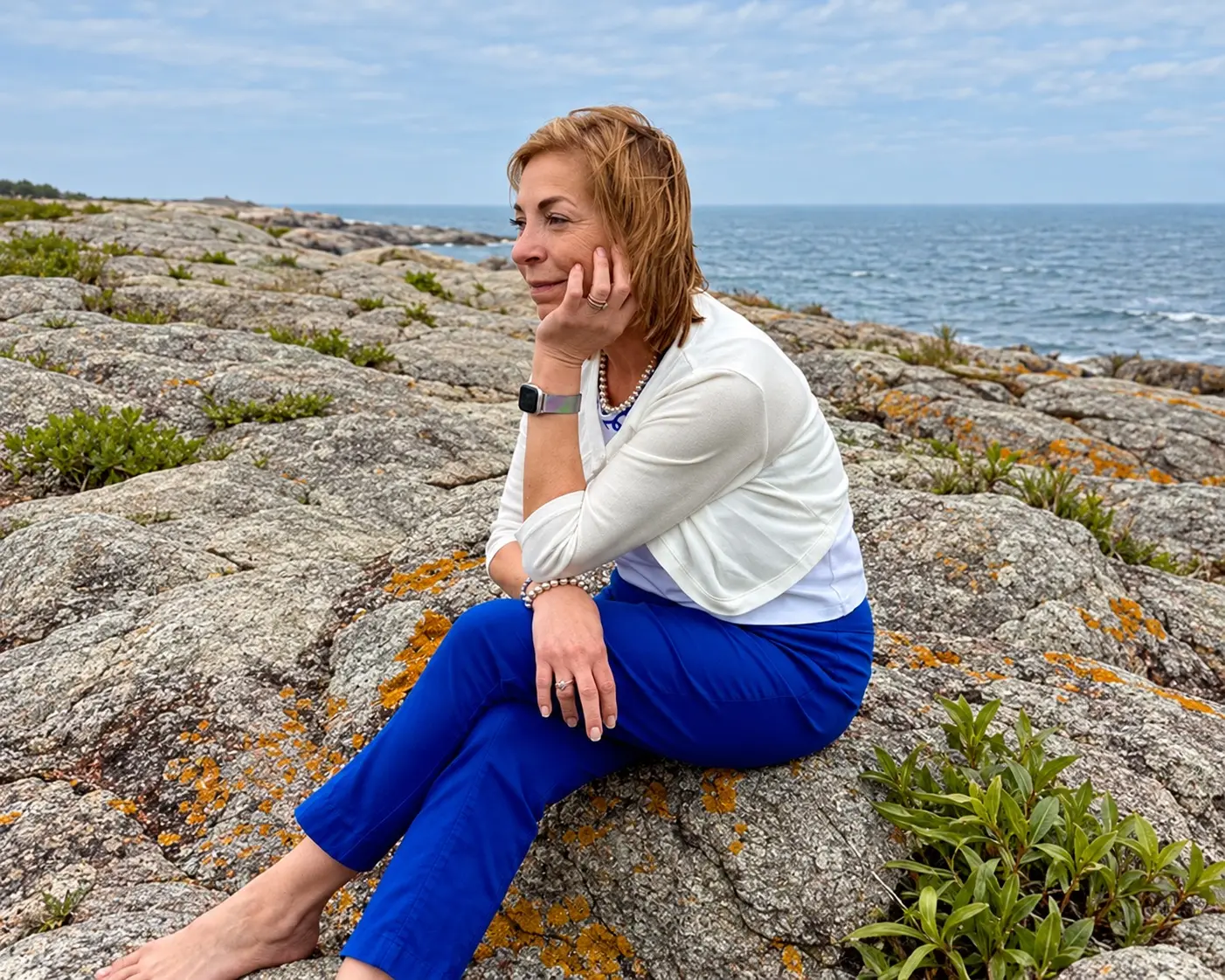 Cynthia Delfino Birdsall sits on coastal rocks with her chin resting on her hand.